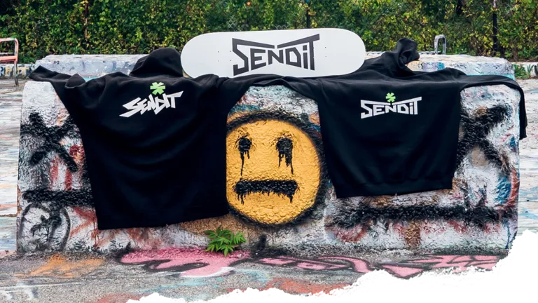 Send It sweatshirts and a skateboard laying on a concrete block at a skate park with a yellow smiley face graffiti surrounding